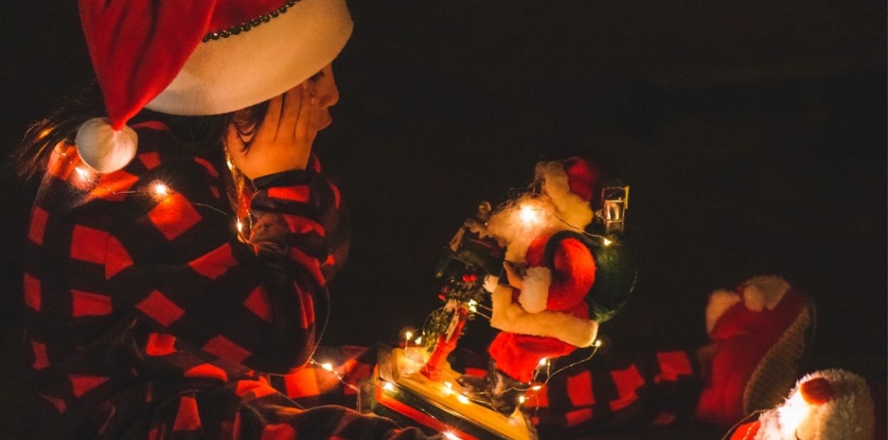A young boy sitting in the dark with his legs out in front of him. He has a Santa Claus figure in his lap and holding his cheeks with his hands in disbelief at the lit up figurine. 